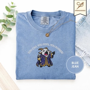 May include: A light blue t-shirt with the embroidered phrase "YOU CAD, YOU FOOL, YOU RUINER!" and a cartoon goat character. The shirt has a crew neck and short sleeves. A gold necklace is also visible.