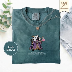 May include: A blue spruce t-shirt featuring an embroidered graphic of a goat wearing a purple wizard robe and holding a pink potion bottle. Below the goat, the text reads: BIANCA PREPARE YOURSELF FOR BYATTLE. The shirt has a crew neckline.