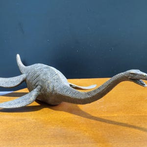 Puede incluir: Una figura de color bronce de un plesiosaurio con cuello largo, cabeza pequeña con la boca abierta y cuatro aletas. La criatura tiene una superficie texturizada y escamosa en todo el cuerpo y está colocada horizontalmente mirando a la derecha.