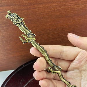Puede incluir: Una figura de metal larga y delgada de color oro antiguo con forma de dragón. El dragón presenta escamas detalladas a lo largo de su cuerpo alargado, una cabeza con cuernos y cuatro patas con garras. La cola termina en un diseño de llama.