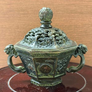 May include: A hexagonal bronze incense burner featuring a dark patina, intricate relief carvings of clouds and circular motifs, two lion-shaped side handles, and a removable lid with an openwork design topped by a spherical finial.