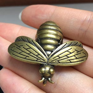 May include: A detailed, antique brass-colored bee-shaped brooch. The bee has textured wings and body segments, with small antennae and legs. The brooch is held in a hand, showcasing its intricate design and metallic finish.