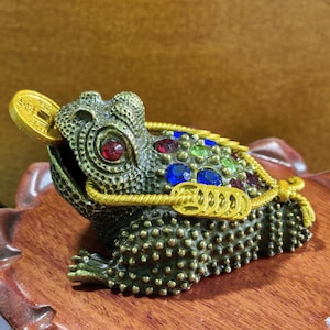 May include: A textured bronze-colored toad figurine featuring a gold-toned coin in its mouth and a string of gold-toned coins draped across its back. The toad is adorned with embedded red, blue, and green faceted stones along its spine.
