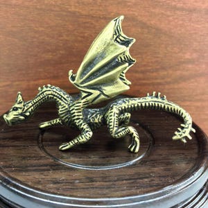 May include: A detailed brass dragon figurine with outstretched wings. The dragon is posed on a dark wooden base. The dragon's scales and wings have intricate detailing, and the overall color is a muted gold.