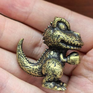 May include: A small, detailed brass figurine of a cartoon Tyrannosaurus Rex. The dinosaur has a textured surface, spiky dorsal plates, and is holding its arms up as if boxing. The figurine is a golden-brown color.