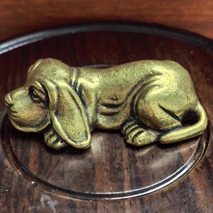 May include: A brass figurine of a basset hound dog in a resting position. The dog has long ears and a detailed face. The figurine is set on a dark wooden surface with a circular design.