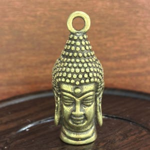 Op de afbeelding: Een kleine, gedetailleerde messing Buddha-hoofdhanger. De hanger heeft een serene uitdrukking, een getextureerde kop en een lus om op te hangen. Het gezicht van de Boeddha is goudkleurig.