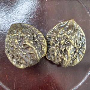 May include: Two metallic, gold-toned objects shaped like walnuts, featuring highly detailed, textured, and irregular surfaces that mimic the natural ridges and crevices of real walnut shells. The objects have a reflective, polished finish.