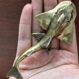 Peut inclure: Une figurine en métal de couleur laiton en forme de poisson-guitare ou de raie, présentant un corps plat et large, une longue queue fine et des détails texturés sur les nageoires et la queue. L'objet a une finition métallique avec des accents sombres.