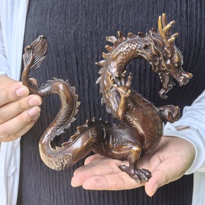 May include: A bronze-colored dragon figurine featuring intricate scale detailing, a curved tail, and a raised claw. The mythical creature is depicted in a dynamic, coiled pose with a textured body, sharp horns, and a detailed face with an open mouth.