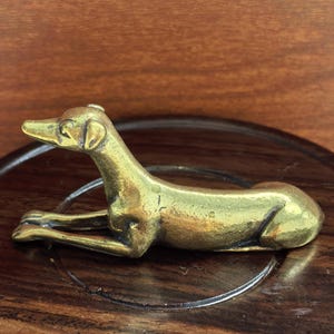 Puede incluir: Pequeña figura de perro de color latón en posición de descanso. El perro está tumbado con las patas delanteras extendidas. La figura está sobre una superficie de madera oscura con un diseño circular. El fondo es de madera.