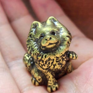 May include: A small, solid brass figurine of a fluffy dog with a thick mane and pointed ears. The metal has a polished gold-toned finish with dark, recessed areas that highlight the textured fur details and the dog's facial features.