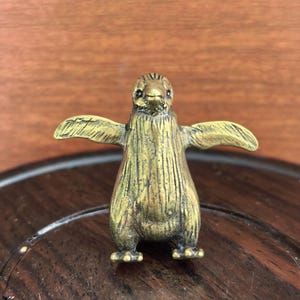 May include: A small, brass penguin figurine with outstretched wings. The penguin has a textured surface and is standing on a dark wooden surface. The figurine is a detailed representation of a penguin.
