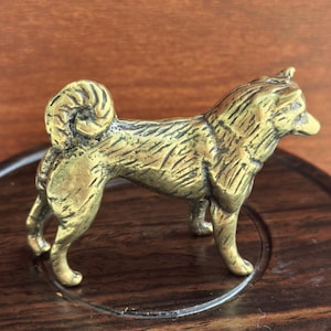 May include: A small, metallic, gold-toned figurine of a dog standing in profile. The sculpture features detailed, textured fur markings, pointed ears, and a curled tail. The dog is depicted in a standing position with its head turned slightly forward.