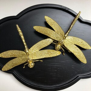 May include: Two gold-coloured dragonfly sculptures with detailed wings rest on a black, cloud-shaped wooden tray. The dragonflies have textured wings and bodies, and are positioned to face opposite directions. The tray has a smooth, matte finish.