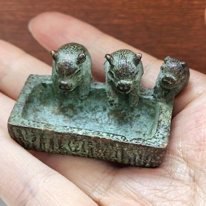 May include: A small, rectangular, weathered bronze-toned metal trough featuring three sculpted pig figures attached to the rim. The object has a textured, oxidized green and brown patina finish, with the three pigs positioned along the edge.