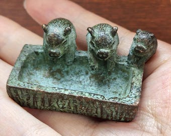 May include: A small, rectangular, weathered bronze-toned metal trough featuring three sculpted pig figures attached to the rim. The object has a textured, oxidized green and brown patina finish, with the three pigs positioned along the edge.