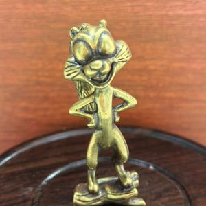 May include: A small, brass-colored figurine of a cartoon squirrel with a smiling face and arms akimbo. The squirrel stands on a small log base. The figurine has detailed features and a vintage aesthetic.