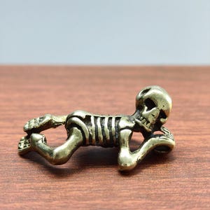 May include: A small, brass-colored skeleton figurine in a reclining pose. The detailed sculpture features a skull, rib cage, and articulated limbs. The figurine is lying on a wooden surface.