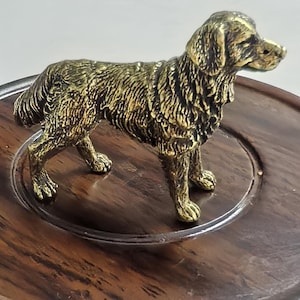 May include: A detailed, golden-colored dog figurine. The sculpture depicts a dog in a standing pose, with textured fur and realistic features. The figurine is placed on a dark wooden surface.