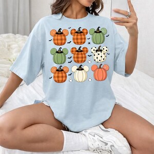 May include: A light blue t-shirt featuring a graphic print of nine pumpkin shapes with mouse-like ears. The pumpkins display various patterns including orange plaid, solid green, white with black polka dots, and solid orange, all wearing small black hats.
