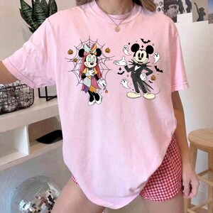 以下が含まれることがあります： ライトピンクのTシャツ。前面には、クモの巣の前に立つパッチワークドレス姿のミニーマウスと、小さな幽霊、コウモリ、オレンジ色のカボチャに囲まれたピンストライプのスーツを着たミッキーマウスのグラフィックプリントが施されています。