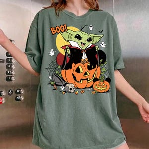 May include: A sage green t-shirt featuring a graphic of a green creature dressed as a vampire sitting on a large orange jack-o'-lantern. The design includes the text BOO!, small white ghosts, a skull, candy corn, and spider webs in the background.