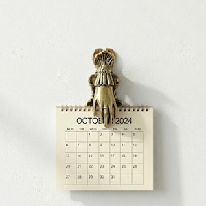 Puede incluir: Un clip de metal color latón con forma de mano y puño con volantes, que sujeta un calendario de papel color crema para octubre de 2024. El calendario presenta una cuadrícula con los días del mes impresos en tinta negra sobre fondo liso.