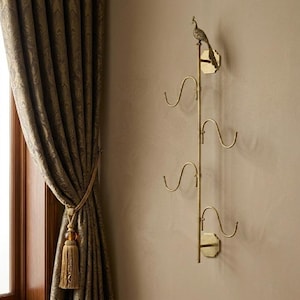 May include: A vertical brass wall-mounted rack featuring a central rod with four curved hooks extending outward. The top of the rod is topped with a decorative peacock figurine. The rack is secured to the wall with two octagonal brass plates.