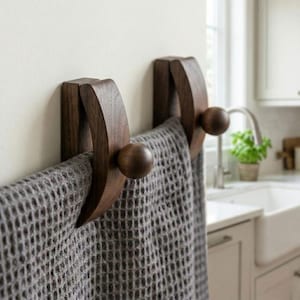 May include: Two dark brown wooden towel hooks mounted on a white wall. Each hook has a curved design with a rounded knob. A gray waffle-weave towel hangs from the hooks. The hooks are in a kitchen setting.
