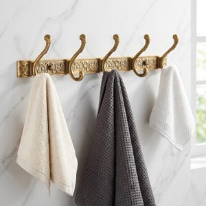 May include: A brass wall-mounted hook rack with five decorative hooks. Three towels in cream, grey, and white hang from the hooks. The rack is mounted on a white marble-patterned wall.