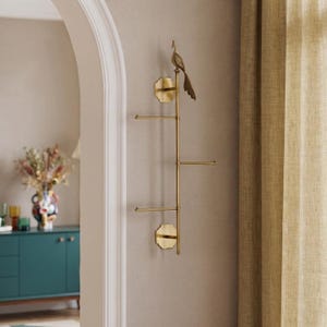 May include: A vertical brass wall-mounted rack featuring a central rod with three horizontal arms extending to the sides. The top of the rack is adorned with a detailed brass peacock figurine. The structure is secured to the wall by two octagonal plates.