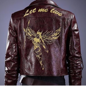 Peut inclure: Une veste en cuir bordeaux foncé avec une broderie dorée représentant une figure ailée tenant une lance au dos. Le texte "Let me live" est brodé en doré au-dessus de la figure. La veste présente un col rabattu.