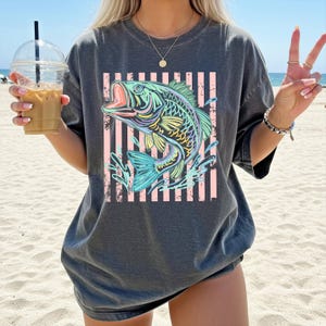 Puede incluir: Camiseta gris oscuro con una ilustraci&oacute;n colorida de un pez con la boca abierta, sobre un fondo de rayas rosas y blancas. El pez es en tonos de azul, verde, amarillo y morado. La camiseta es de estilo casual.