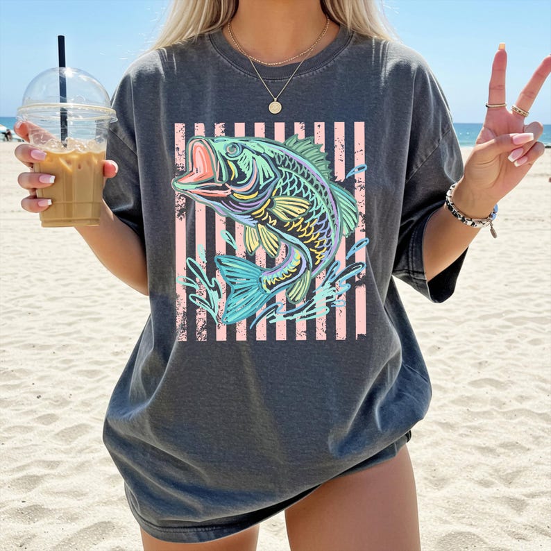 Puede incluir: Camiseta gris oscuro con una ilustraci&oacute;n colorida de un pez con la boca abierta, sobre un fondo de rayas rosas y blancas. El pez es en tonos de azul, verde, amarillo y morado. La camiseta es de estilo casual.