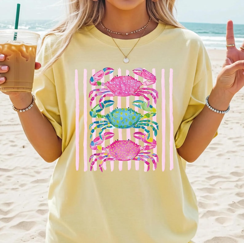 May include: Yellow t-shirt featuring a watercolor design of three colorful crabs stacked vertically. The crabs are pink, blue, and green, set against a backdrop of pink vertical stripes. The shirt is a casual, beach-themed apparel item.
