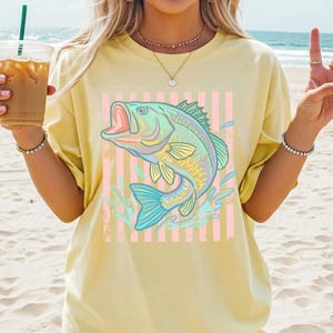 Puede incluir: Camiseta amarilla clara con un dise&ntilde;o de pez colorido. El pez, en tonos azules y verdes, se representa con la boca abierta, sobre un fondo de rayas rosas y blancas. Salpicaduras de agua rodean el pez. Un estilo casual y c&oacute;modo.