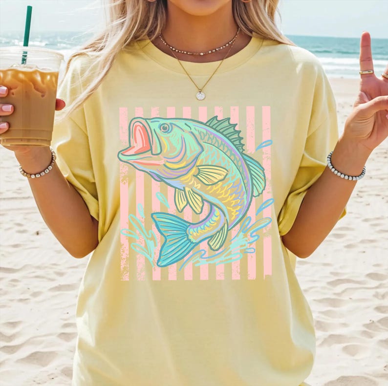 Puede incluir: Camiseta amarilla clara con un dise&ntilde;o de pez colorido. El pez, en tonos azules y verdes, se representa con la boca abierta, sobre un fondo de rayas rosas y blancas. Salpicaduras de agua rodean el pez. Un estilo casual y c&oacute;modo.