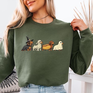 May include: An olive green crewneck sweatshirt featuring a graphic print of four dachshunds in Halloween costumes. The dogs are dressed as a witch with a black hat and cape, a mummy wrapped in white bandages, a hot dog, and a ghost under a white sheet.