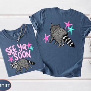 May include: Two denim blue t-shirts with raccoon graphics and the text "See Ya Soon". One shirt features the raccoon walking with pink and teal stars, while the other shows the raccoon from behind with stars. The shirts have a casual, relaxed fit.
