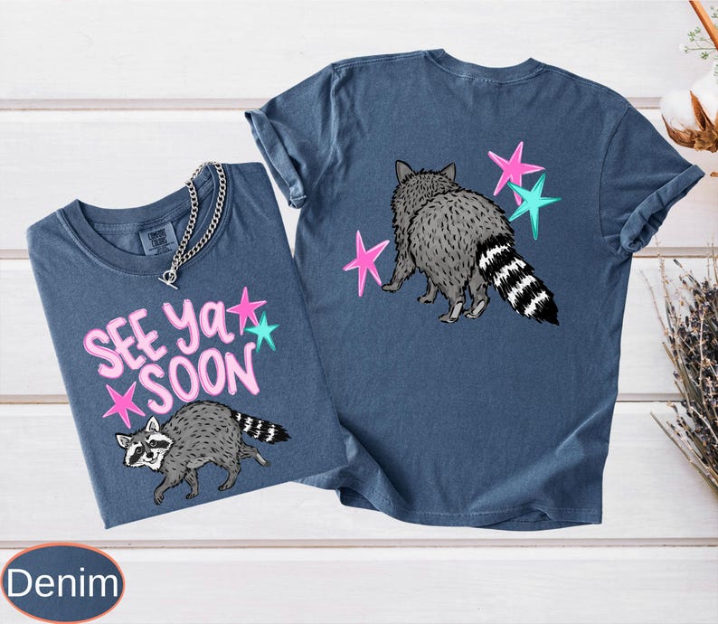May include: Two denim blue t-shirts with raccoon graphics and the text "See Ya Soon". One shirt features the raccoon walking with pink and teal stars, while the other shows the raccoon from behind with stars. The shirts have a casual, relaxed fit.