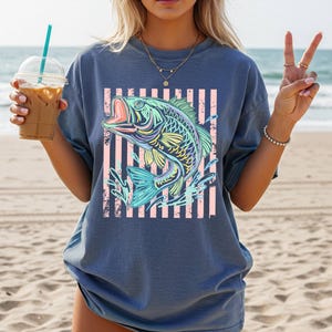 Puede incluir: Una camiseta azul con un gr&aacute;fico colorido de un pez con la boca abierta, sobre un fondo de rayas rosas. El dise&ntilde;o del pez incorpora tonos de azul, amarillo y verde, creando una imagen vibrante y llamativa.
