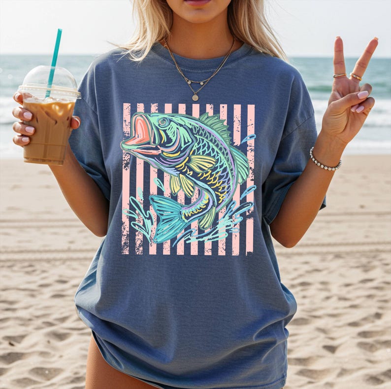 Puede incluir: Una camiseta azul con un gr&aacute;fico colorido de un pez con la boca abierta, sobre un fondo de rayas rosas. El dise&ntilde;o del pez incorpora tonos de azul, amarillo y verde, creando una imagen vibrante y llamativa.