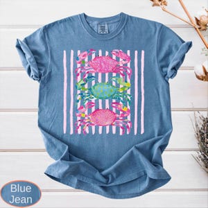 May include: Blue jean colored t-shirt with a watercolor design of three crabs. The crabs are pink and blue, set against a background of pink vertical stripes. The shirt is a casual, comfortable style.