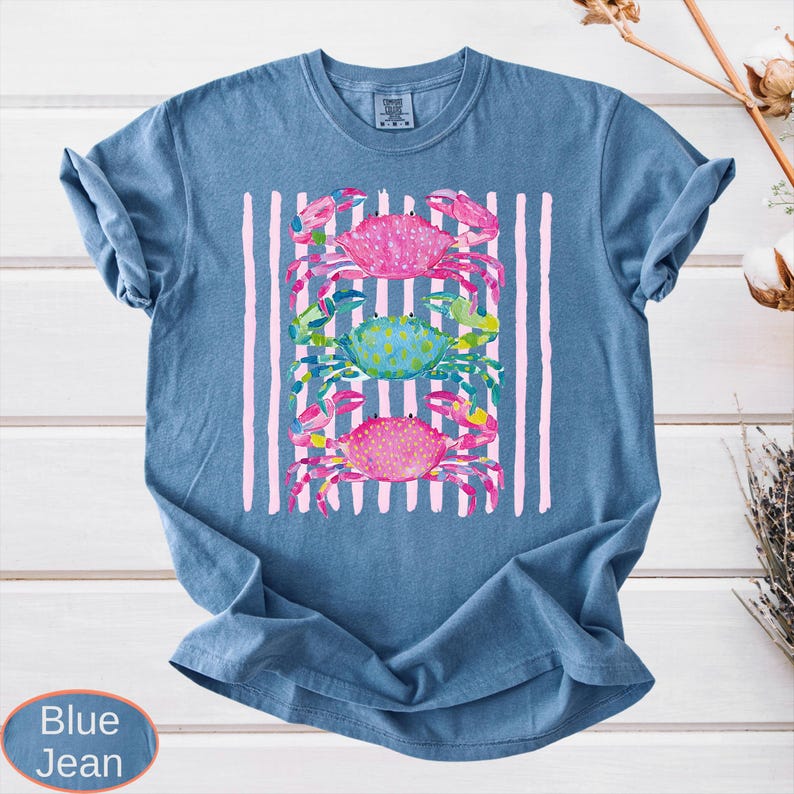 May include: Blue jean colored t-shirt with a watercolor design of three crabs. The crabs are pink and blue, set against a background of pink vertical stripes. The shirt is a casual, comfortable style.