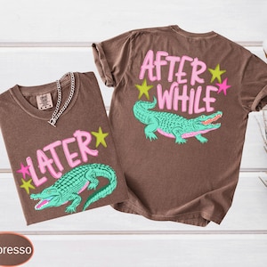 Puede incluir: Dos camisetas color espresso con dise&ntilde;os a juego. Una camiseta presenta el texto "LATER" y la otra "AFTER WHILE", ambas en rosa, con un caim&aacute;n verde y detalles de estrellas. Una cadena de plata est&aacute; en una camiseta.