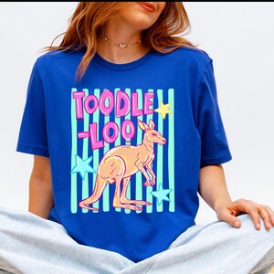May include: Royal blue t-shirt featuring a cartoon kangaroo with the text "TOODLE LOO" in pink and light blue. The design includes light blue vertical stripes and star accents in yellow and blue.