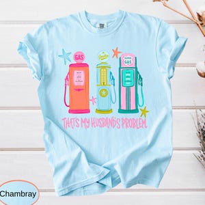 May include: Light blue t-shirt featuring three vintage gas pumps in coral, yellow, and teal, with star accents. The text "THAT'S MY HUSBAND'S PROBLEM" is printed in pink below the pumps. The shirt is made of chambray fabric.
