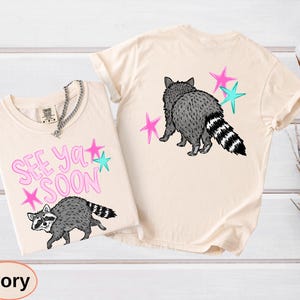 May include: Ivory-colored t-shirts with raccoon graphics. One shirt features the text "SEE YA SOON" with pink and teal stars. The other shirt shows a raccoon with pink and teal stars.
