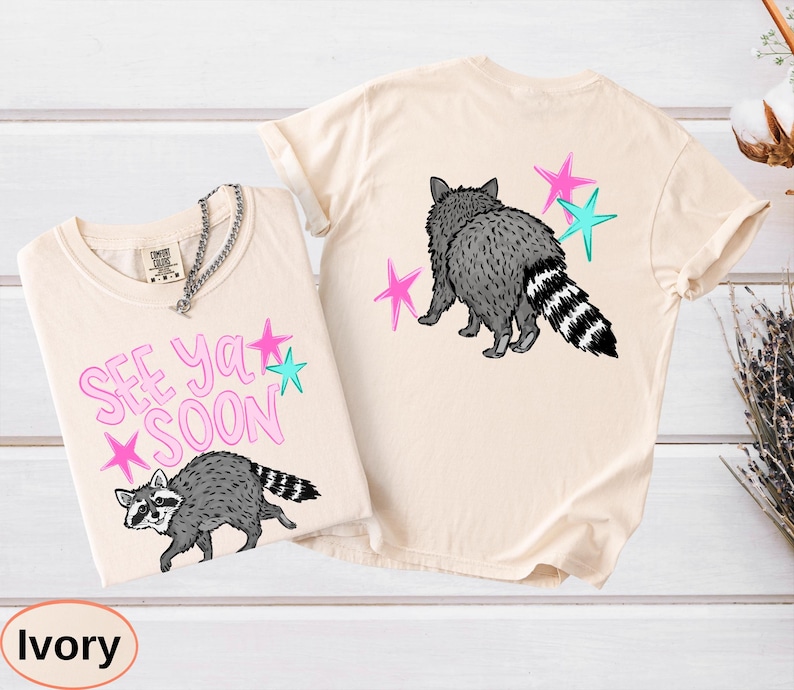 May include: Ivory-colored t-shirts with raccoon graphics. One shirt features the text "SEE YA SOON" with pink and teal stars. The other shirt shows a raccoon with pink and teal stars.